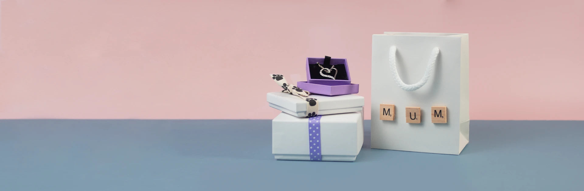 Gift box, jewelry, and 'MUM' sign on a blue surface with pink background