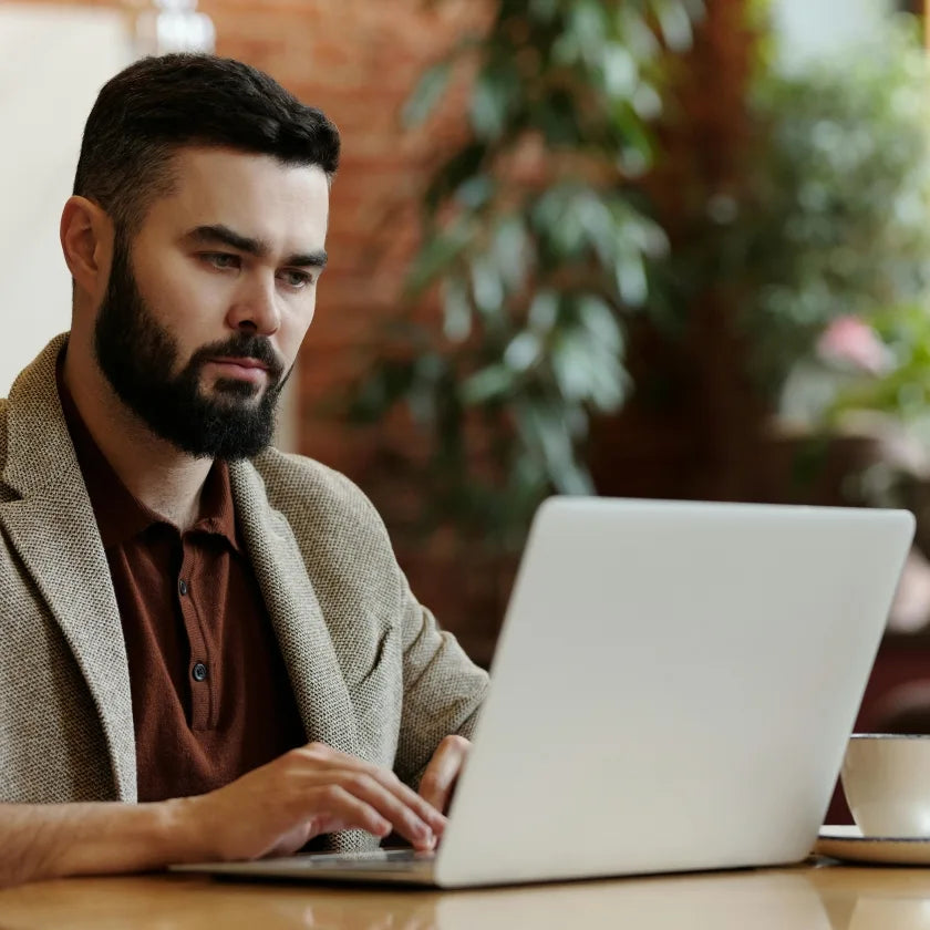 A bearded man sits at a wooden table, intently focused on his laptop. He wears a brown shirt and beige blazer in a cozy, plant-filled room.