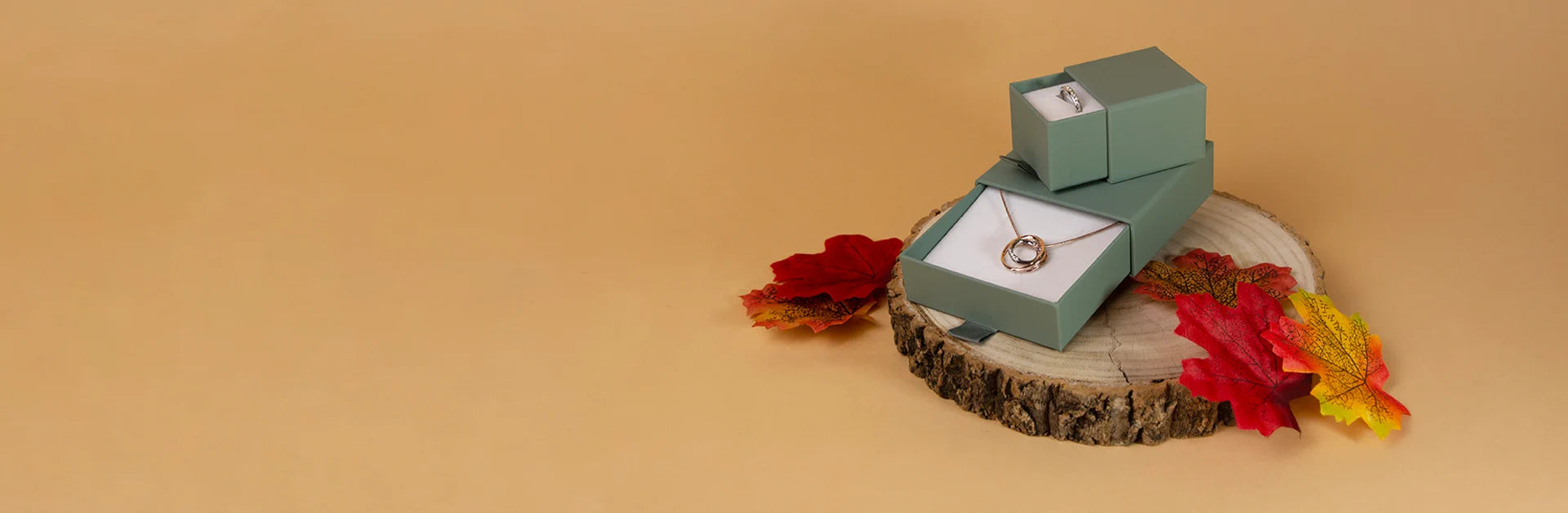 Two mint green jewelry gift boxes stacked on a rustic wood slice, with a gold ring displayed on the front box. Red and yellow autumn leaves are placed around the stack against a warm beige background.