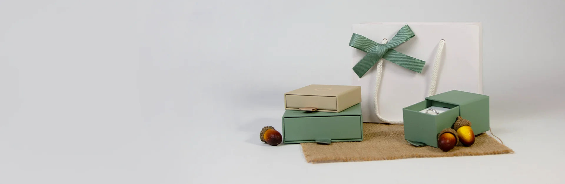 Gift boxes and a white shopping bag with a green bow are arranged on burlap, alongside acorns and chestnuts, against a light background.