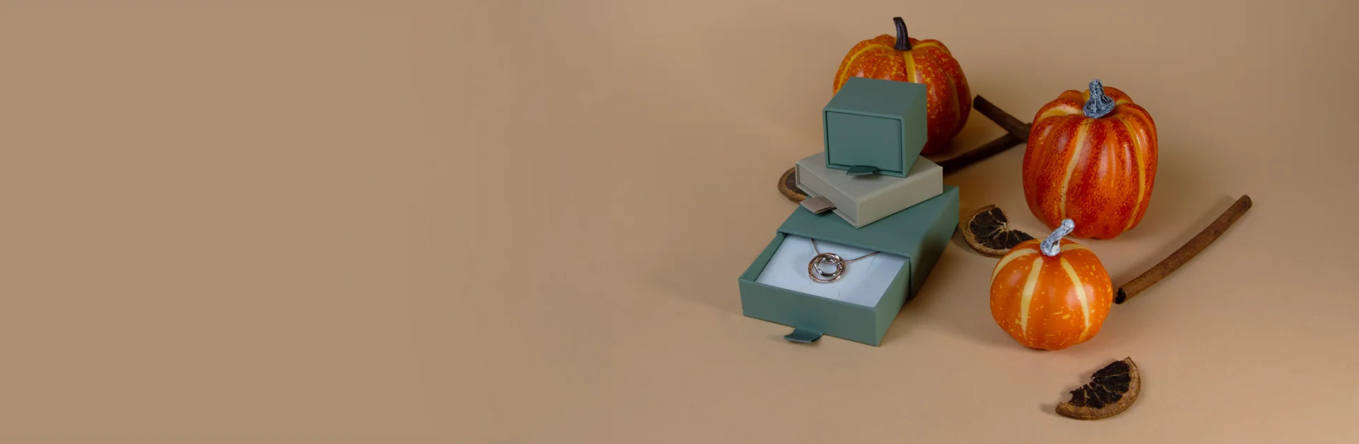 A necklace in a stack of small gift boxes, arranged with three decorative pumpkins, cinnamon sticks, and dried orange slices on a tan background.