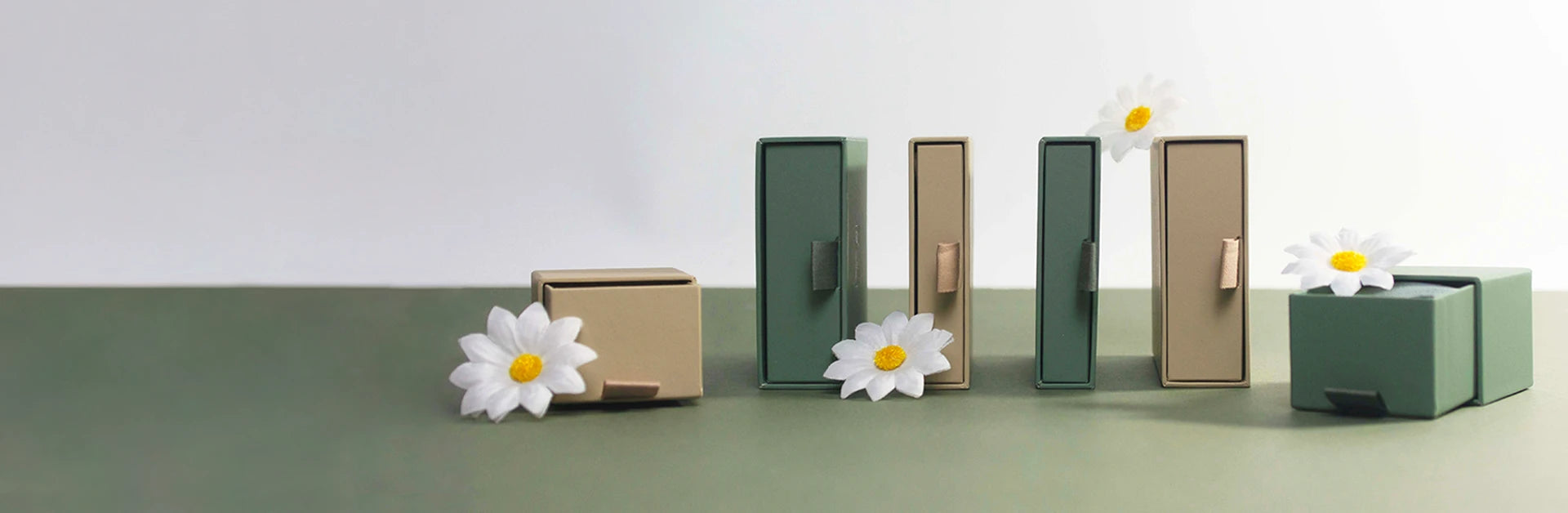 A set of small minimalist storage boxes in muted green, beige, and brown tones, arranged on a two-tone surface with white daisy flowers placed around them.