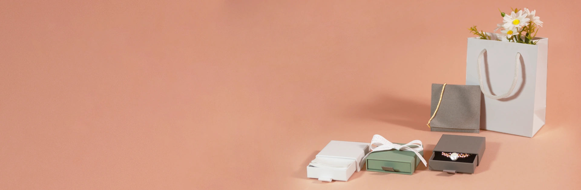 A styled flat-lay of gift packaging on a solid peach background, featuring a white paper shopping bag with a gold ribbon and daisies, a gray gift box, a green box, and a white box with a white ribbon bow.
