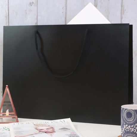 A large black gift bag with a black handle sits on a white surface against a light wooden wall. A corner of white paper peeks from the bag. In the foreground, there's a magazine, a geometric candle holder, and a blue patterned cup, creating a cozy, stylish ambiance.
