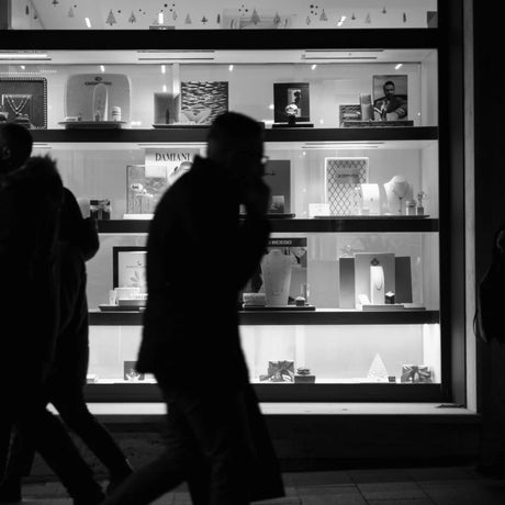 Silhouetted pedestrians pass by a brightly lit store window displaying jewellery and decorative items, creating a contrast in a nighttime urban scene.