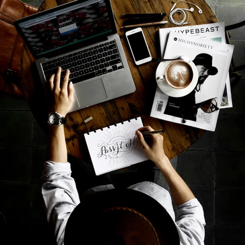 Person working at a desk with a laptop, phone, coffee, and magazine.