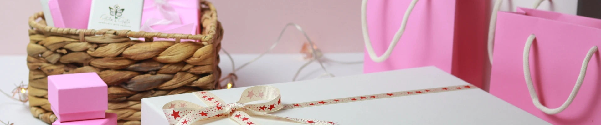 A large white gift box with a star-patterned ribbon sits in front. Surrounded by pink gift bags and a wicker basket full of small, pink gift boxes.