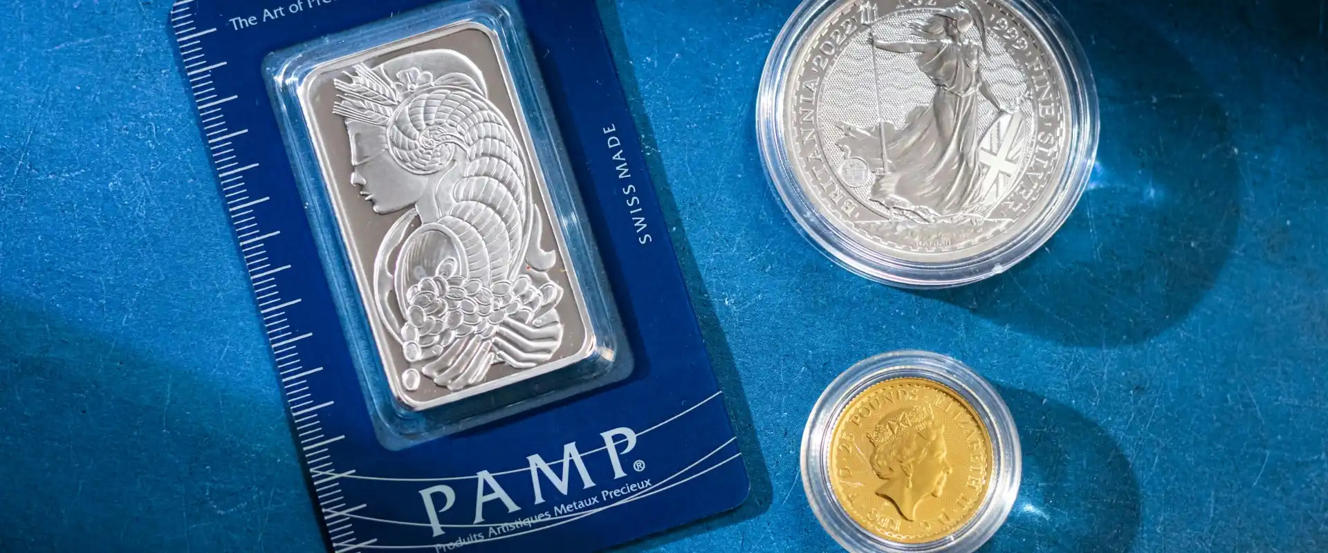Silver and gold coins and a silver bar against a blue background. The bar features intricate designs and is labeled 