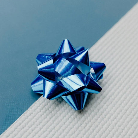 A shiny blue metallic gift bow sits on a textured white surface with a background of blue. The tone is festive and elegant.