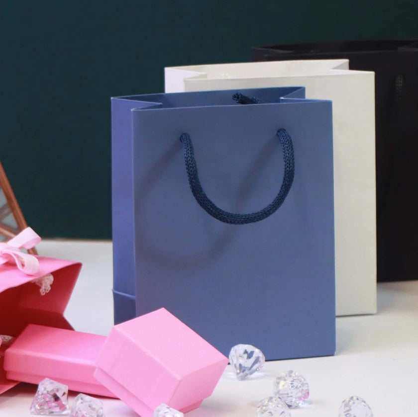 Three gift bags in blue, white, and black stand on a surface adorned with pink boxes and scattered crystal-like gems, conveying a sense of celebration.