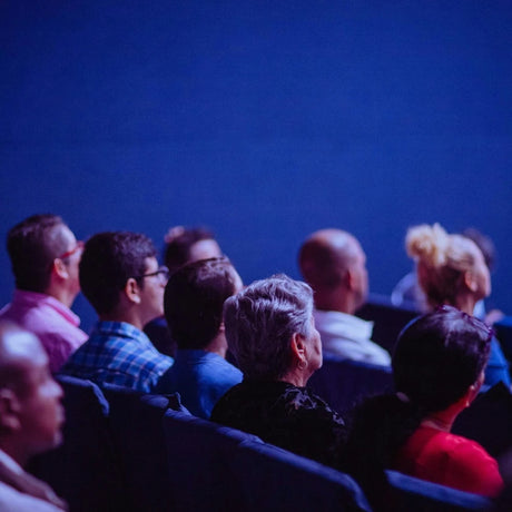 Audience seated in a dark theater, attentively watching an event. The image conveys focus and anticipation, with a calm, immersive atmosphere.