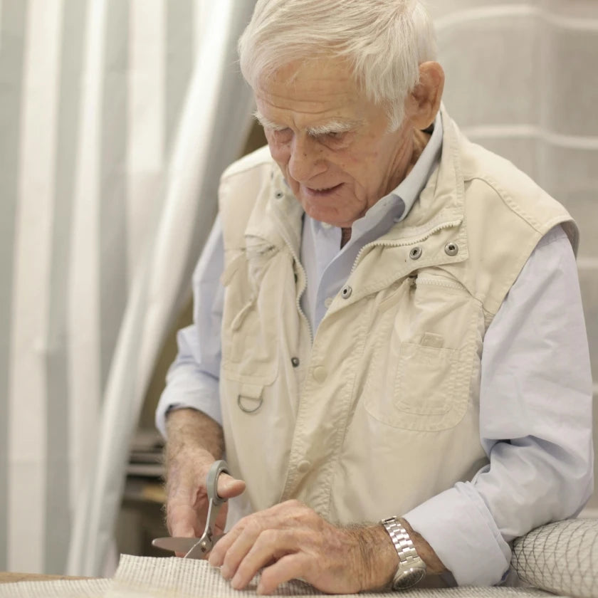An elderly man (likely a small business owner) with white hair. Wearing a jacket vest, blue shirt and a watch. Cutting fabric on a table with a pair of scissors.
