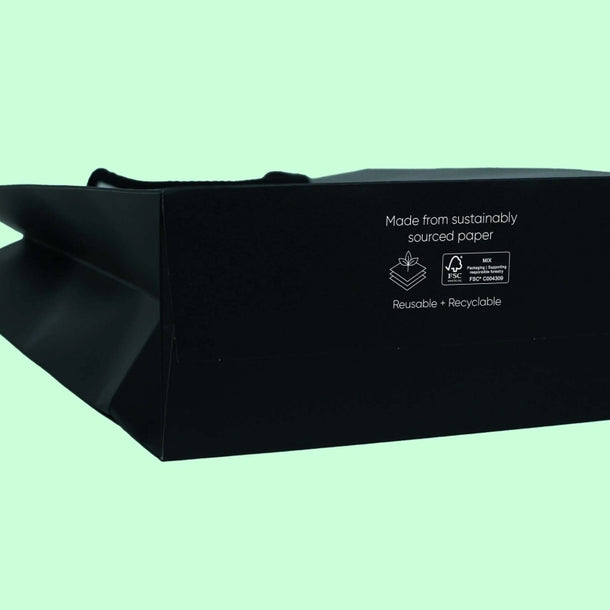 Black paper bag on a light green background. Text reads, "Made from sustainably sourced paper. Reusable + Recyclable." Eco-friendly theme.