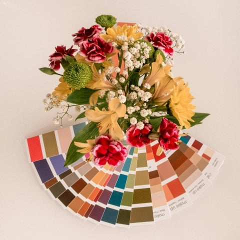 A colorful bouquet with red, yellow, and white flowers sits atop a fan of paint swatches, creating a vibrant, cheerful contrast on a plain background.