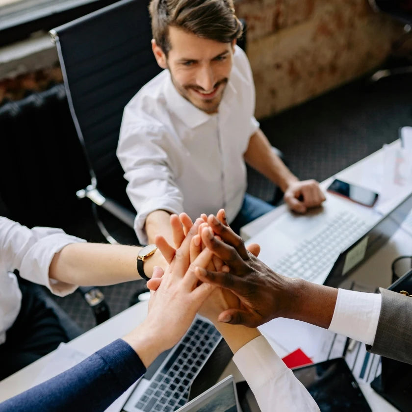 A group of people in business attire gather around a desk, joyfully stacking hands in a teamwork gesture. Laptops and phones are visible, suggesting a collaborative office setting.