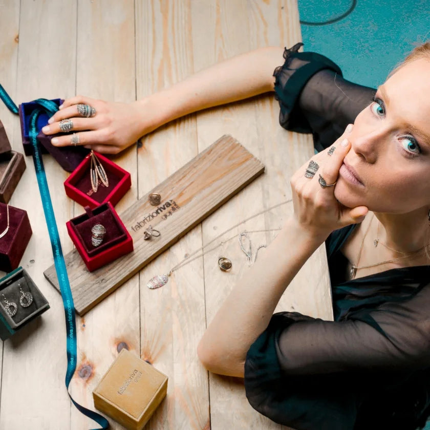 A woman with a thoughtful expression showcases silver rings and earrings on a wooden table. Velvet boxes, a branded wood plank, and a gold box accentuate the luxury feel.
