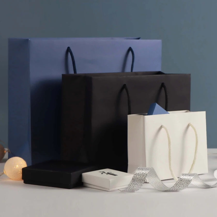 An organised display of elegant kraft gift bags in blue, black, and white, accompanied by gift boxes and a decorative ribbon on a neutral backdrop.