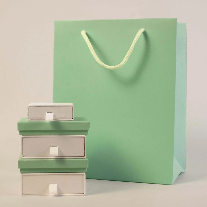 A stack of three small green and white gift boxes sits beside a large mint green shopping bag with rope handles, set against a light grey background.