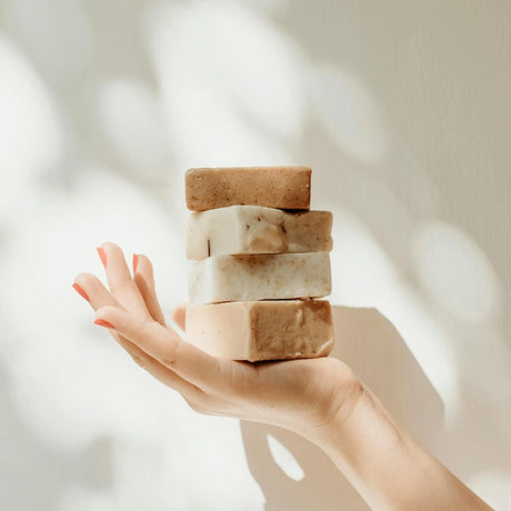 A hand with pink nail polish holds a stack of four rectangular soap bars in neutral tones, set against a soft, sunlit background, conveying calmness.