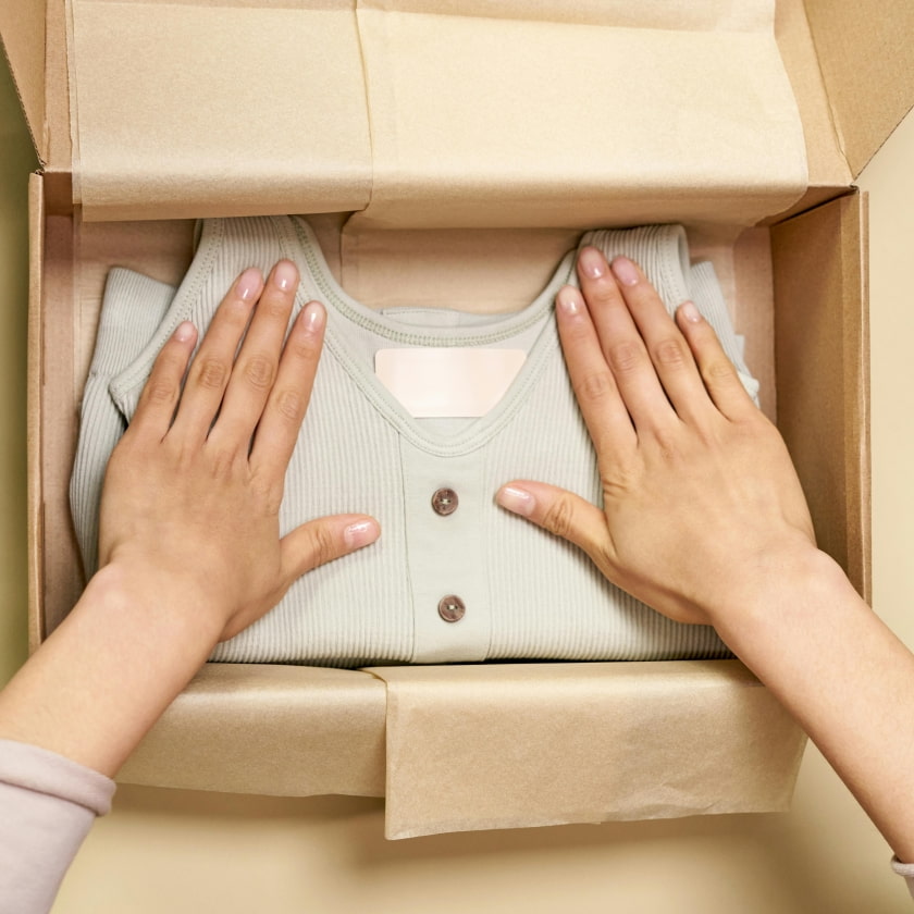 Hands placing a light green ribbed bodysuit with wooden buttons inside an open brown cardboard box with packing paper, on a cream background.
