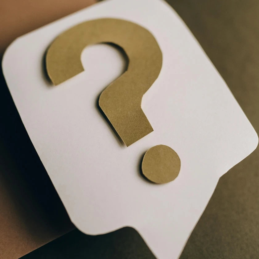 A cut-out paper question mark in brown sits on a white speech bubble cutout, placed on a dark surface. The image conveys curiosity and inquiry.