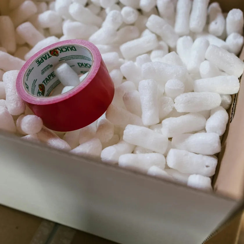 A box containing white plastic pellets alongside a roll of red tape.
