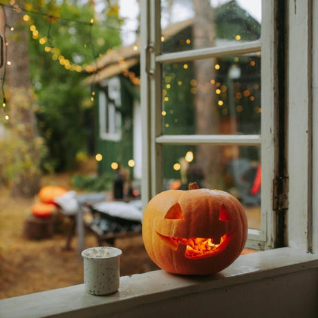 A carved pumpkin glows warmly on a windowsill, beside a candle, with festive string lights and a cozy autumn garden scene in the background.