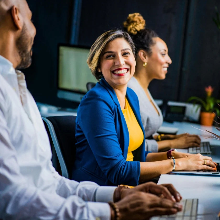 Smiling woman in a blue blazer and yellow top sits at a desk with colleagues, typing on keyboards in a positive, collaborative office setting.