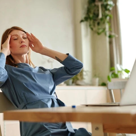 A woman in a blue dress sits at a desk with a laptop, rubbing her temples, eyes closed, appearing stressed. Sunlight filters through a window, highlighting indoor plants.