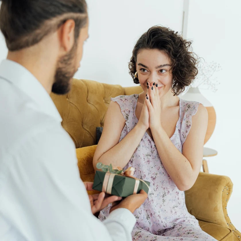 A man surprising a woman with a green gift. The woman has a brown curly bob, and is wearing a purple dress. They are sat on a yellow sofa, with a white background.
