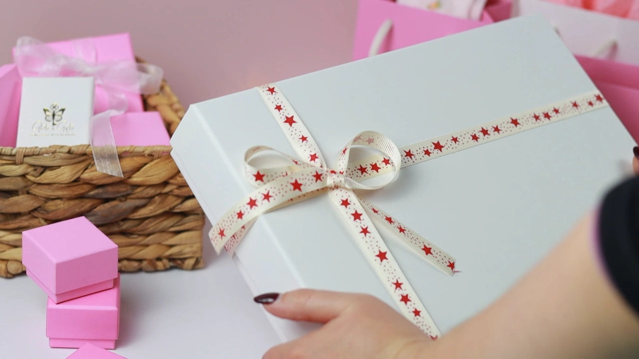 A hand holds a white gift box adorned with a red star ribbon. Nearby, a wicker basket and pink gift boxes exude a celebratory mood.