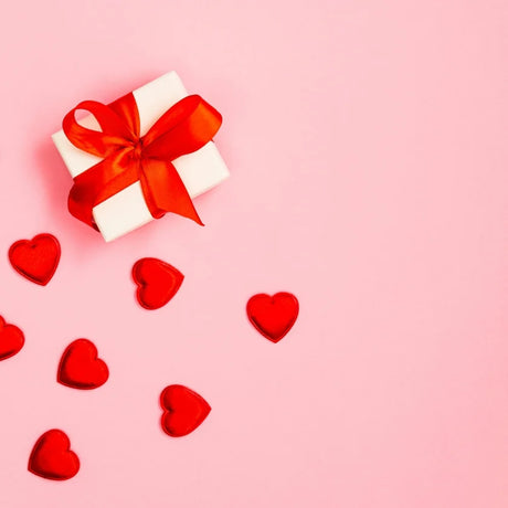 A white gift box with a red ribbon sits on a pink background, surrounded by scattered red heart shapes, conveying a romantic and cheerful tone.