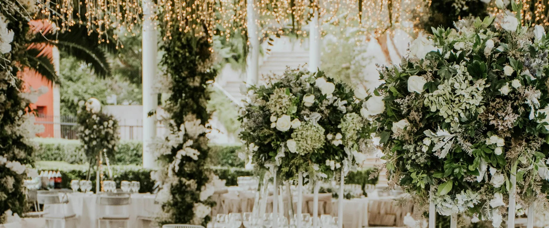 Elegant outdoor wedding setup with white tables and chairs, topped with lush floral centerpieces. Fairy lights hang above, creating a romantic ambiance.
