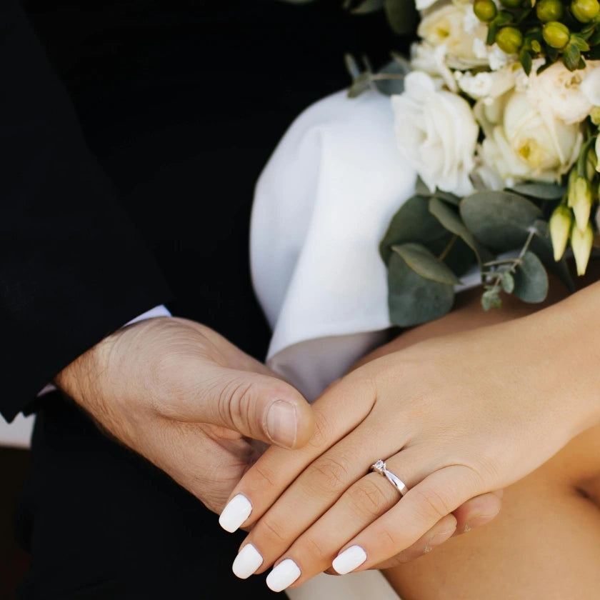 Close-up of a couple holding hands, highlighting a woman's engagement ring. A bouquet of white flowers is visible, conveying a romantic, celebratory mood.