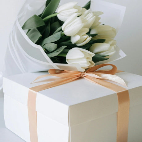 Bouquet of white tulips rests on a white gift box tied with a peach-colored ribbon, conveying elegance and celebration.