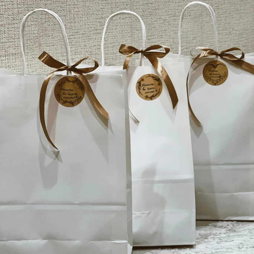 Three white gift bags with brown ribbon bows and round tags. The bags are side by side against a textured background, conveying a festive tone.