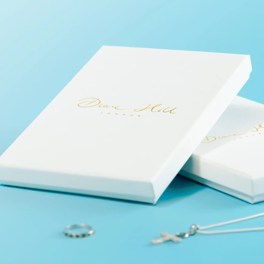 White jewelry boxes with gold script on a light blue background, alongside a simple silver ring and a delicate cross necklace, convey elegance.