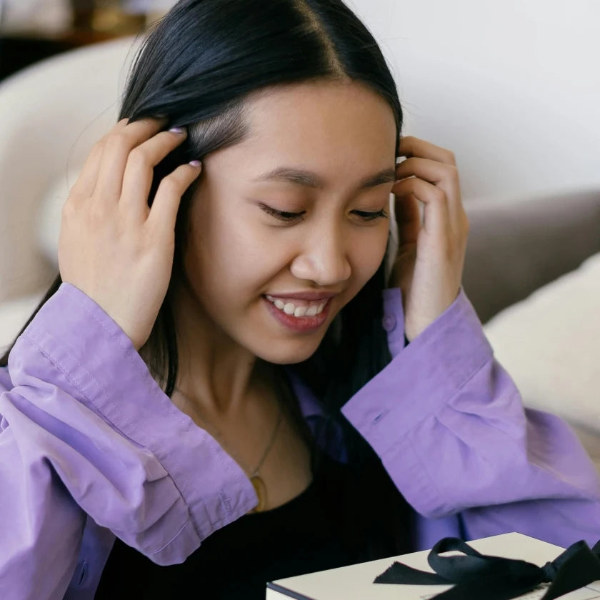 Young woman with long black hair and a purple shirt, smiles while holding her hair back, looking at a gift box with a black ribbon, conveying joy.