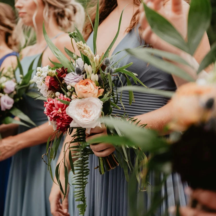 Bridesmaids in blue dresses hold vibrant bouquets with roses, lilies, and greenery. The tone is elegant and joyful, featuring close-up floral details.