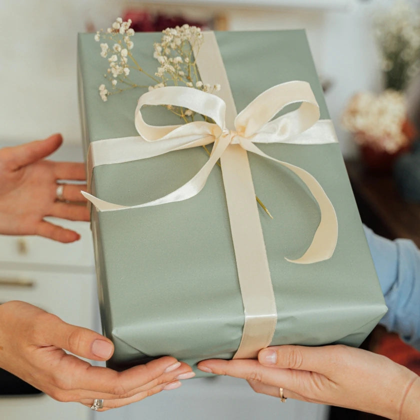 Two people exchanging a green gift box wrapped with a cream ribbon and decorated with delicate flowers, conveying a sense of celebration and goodwill.