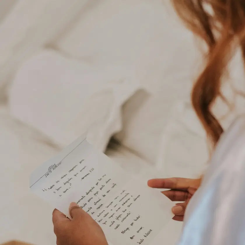 A person with auburn hair reads a handwritten letter on white paper against a blurred, soft-focus background of white sheets, evoking a sense of intimacy and reflection.
