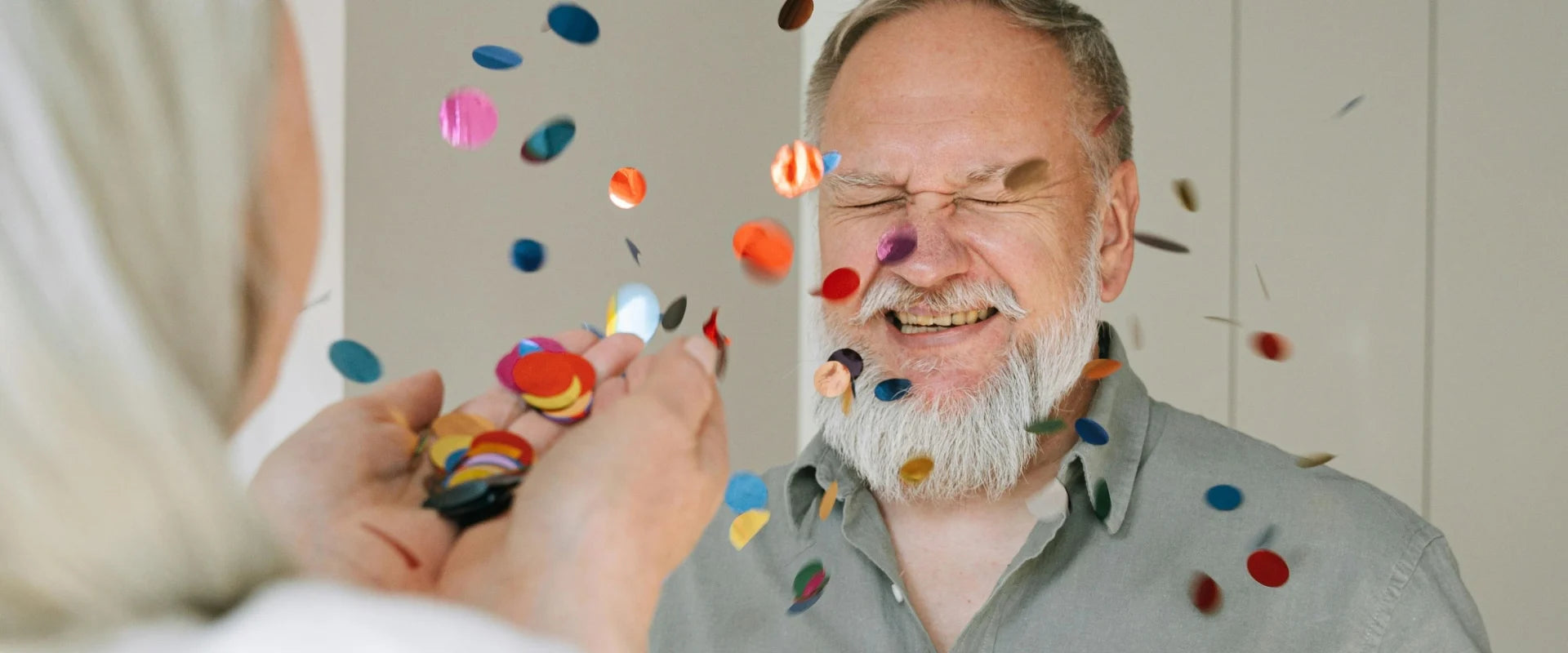 A bearded father with eyes closed smiles joyfully as colorful confetti is thrown at him. The atmosphere is festive and lighthearted.