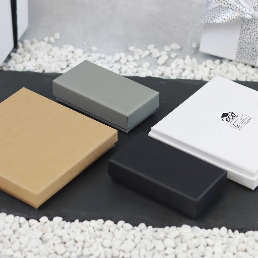 Four rectangular boxes in different colors - grey, black, white with a logo, and brown- are arranged on a black surface, surrounded by white pebbles.