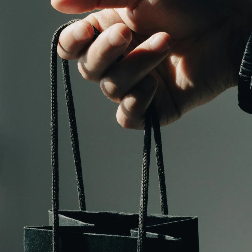 A hand gently holds a small black paper bag by the handles against a soft gray background, suggesting a minimalistic and calm atmosphere.