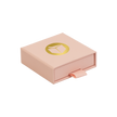 A light pink square gift box with a pull tab and a gold circular logo on top, shown on a plain white background.