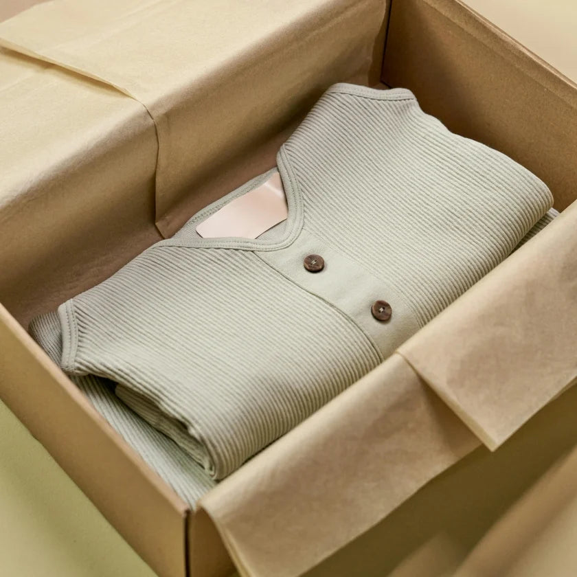 A neatly folded, light green ribbed sweater with two wooden buttons is placed in an open cardboard box lined with beige tissue paper, conveying a sense of elegance and warmth.