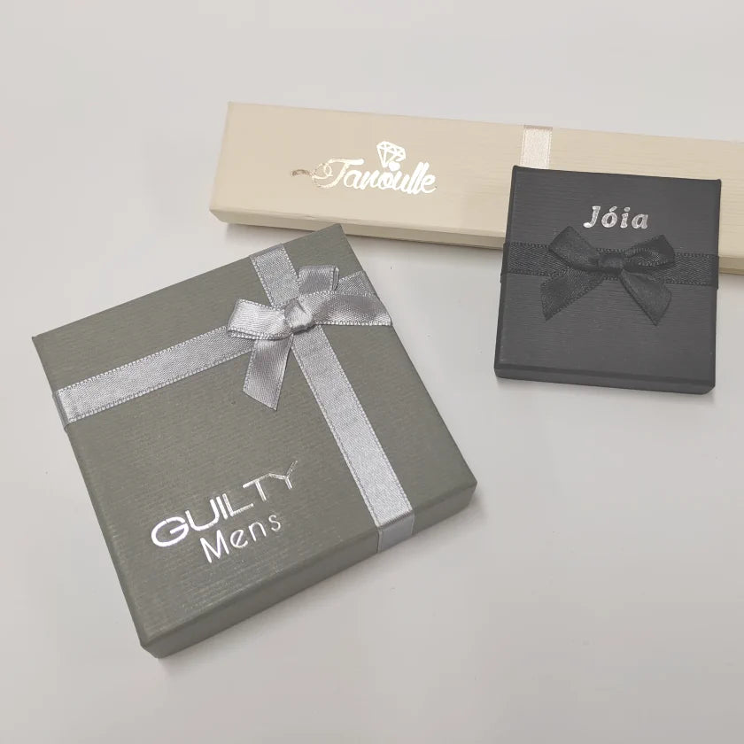 Three elegant gift boxes — a gray GUILTY Men box with a silver ribbon, a cream “Tavolille” box, and a black “Joia” box — arranged on a pale surface, creating a refined, luxurious mood.