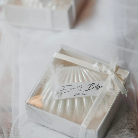 Elegant wedding favour in a clear box, adorned with a white ribbon and label with names and a date. Background features soft tulle fabric.