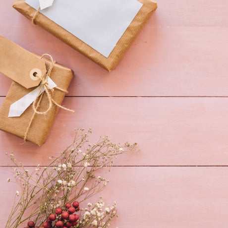 Two brown paper-wrapped gifts with tags lie on a pink wooden surface. Nearby, white flowers and red berries add an elegant touch.
