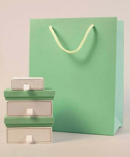 A large mint green gift bag with a white rope handle stands behind three stacked green and white jewellery boxes.